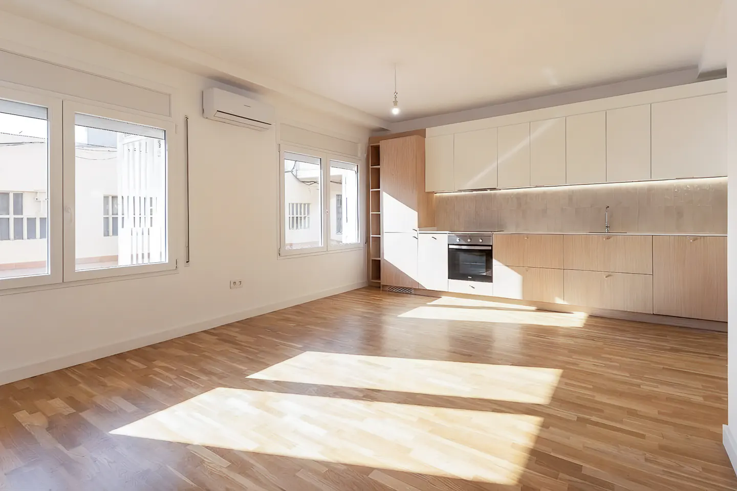 Bright, empty apartment with hardwood floors, white walls, and a modern kitchen with light wood cabinets and stainless steel appliances. Sunlight streams through the windows.