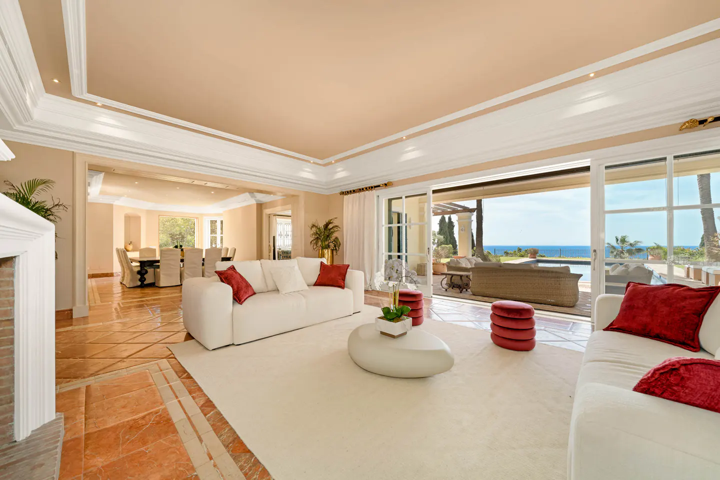 Bright living room with white sofas, red pillows, and a view of the ocean through sliding glass doors.