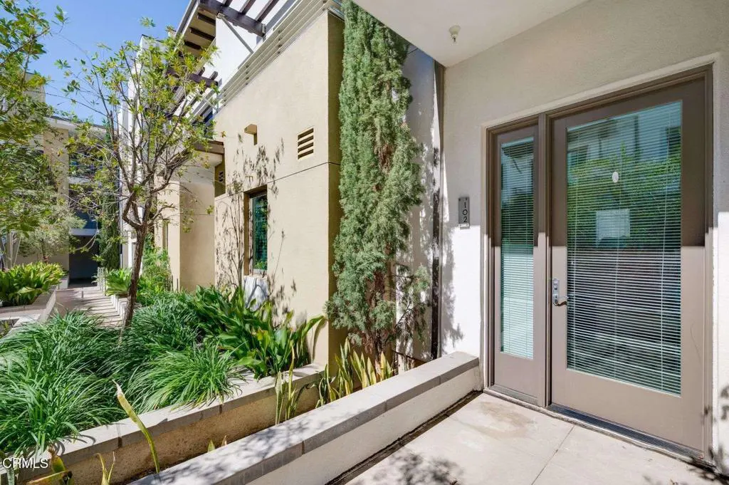 Exterior view of a beige building with a patio, green plants, and a double door with blinds. Unit number 102 is visible.