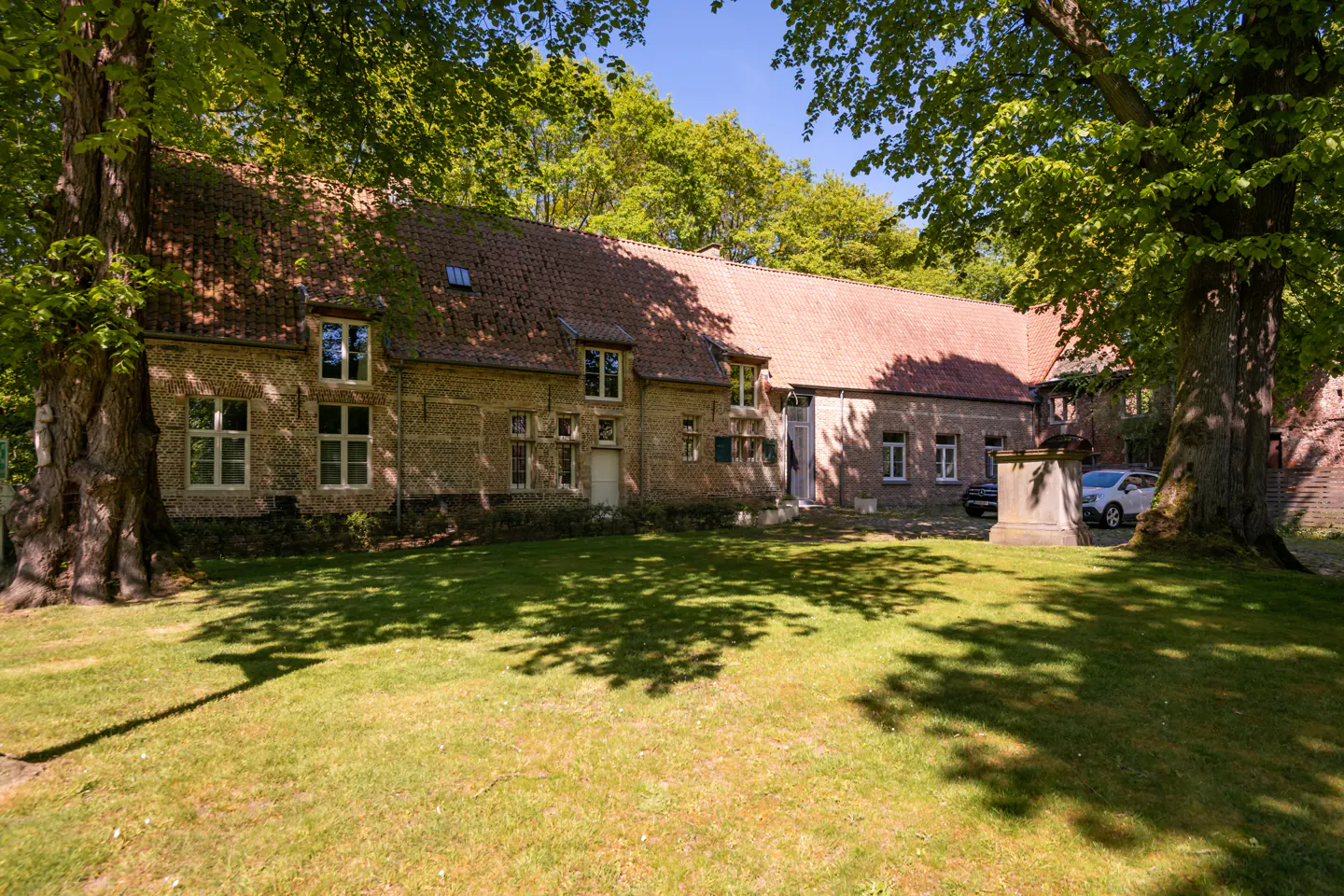 Exterior view of a long, brick house with a red tile roof, surrounded by green trees and a lawn with shadows.