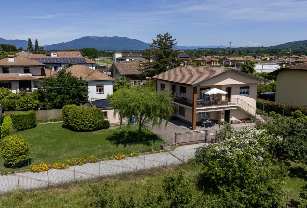 Two-story house with a brown roof, a balcony with a white umbrella, and a green lawn. Mountains are visible in the background.