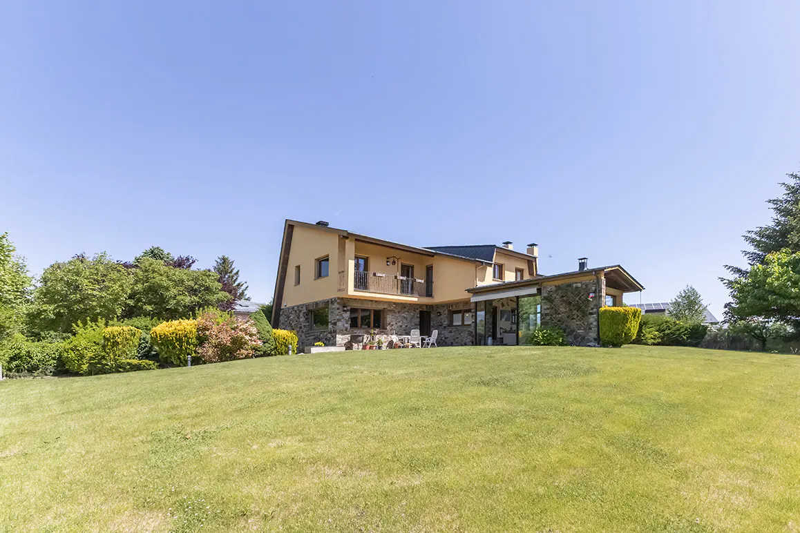 Two-story house with stone and yellow stucco exterior, balcony, and patio, set on a large green lawn under a clear blue sky.