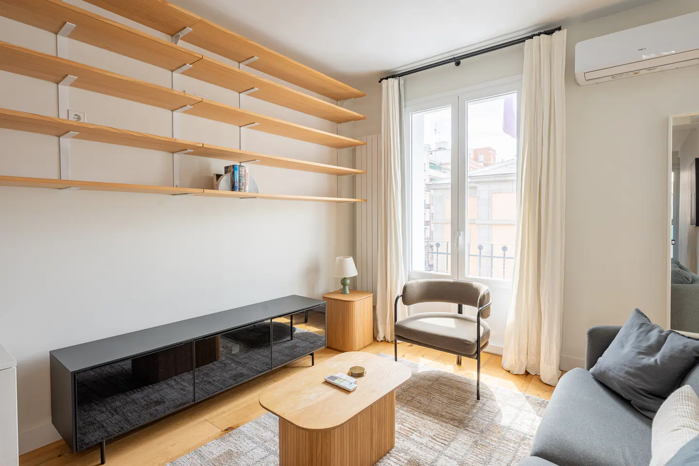 Bright living room with light wood shelves, gray media console, wood coffee table, and gray sofa near a window with white curtains.