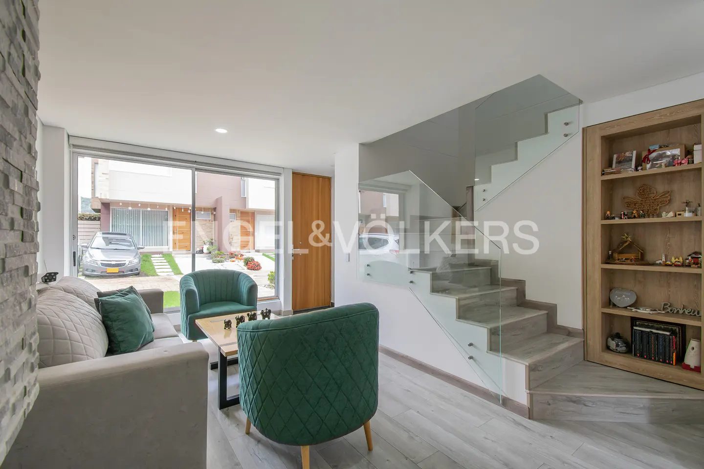 Living room with gray sofa, green chairs, and glass staircase. Outside view of a car and house.