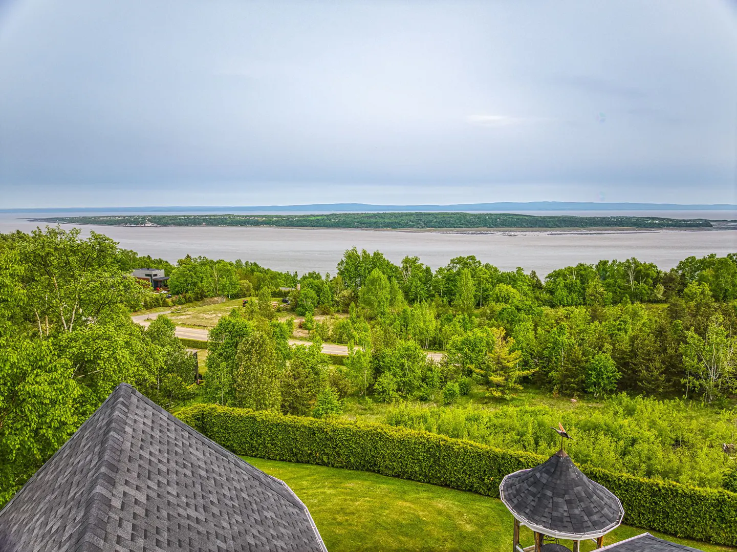 Scenic view from a property overlooking lush green trees, a body of water, and a distant shoreline under a cloudy sky.