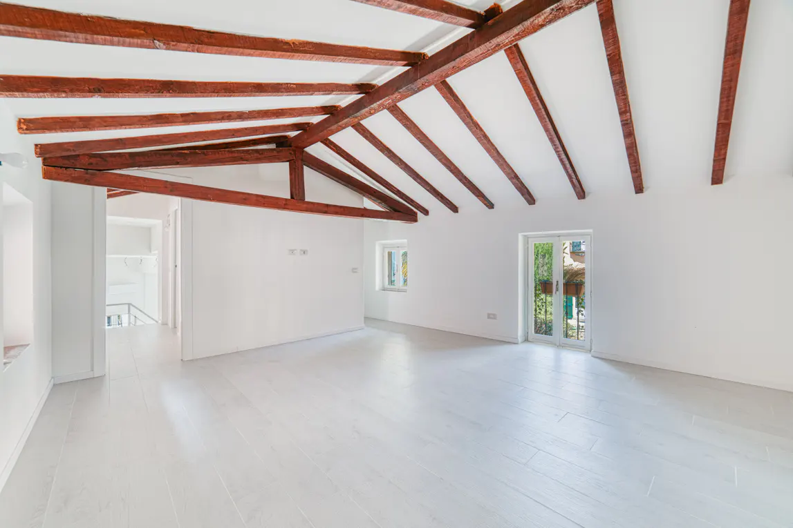 Bright, empty room with white walls, white wood floors, and exposed dark wood ceiling beams. A window and glass doors let in natural light.