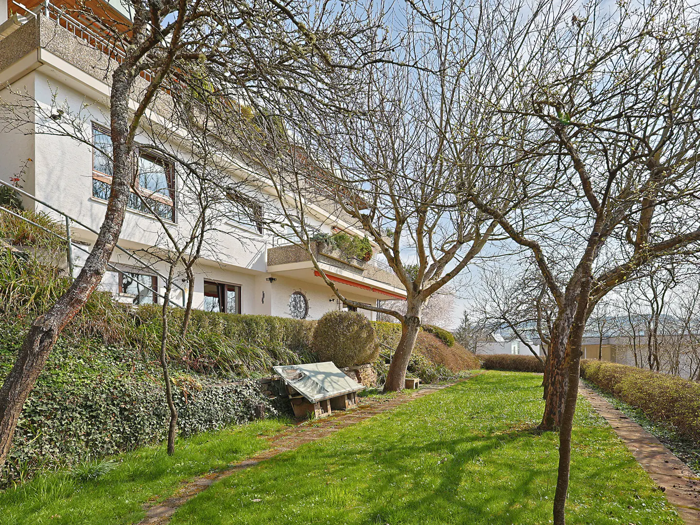 A white two-story house with a balcony, viewed through bare trees and a green lawn.