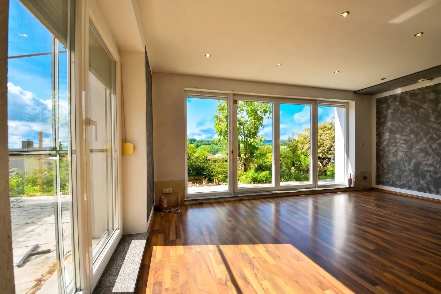 Bright, empty room with hardwood floors and large windows overlooking a green garden. Sunlight shines on the floor.