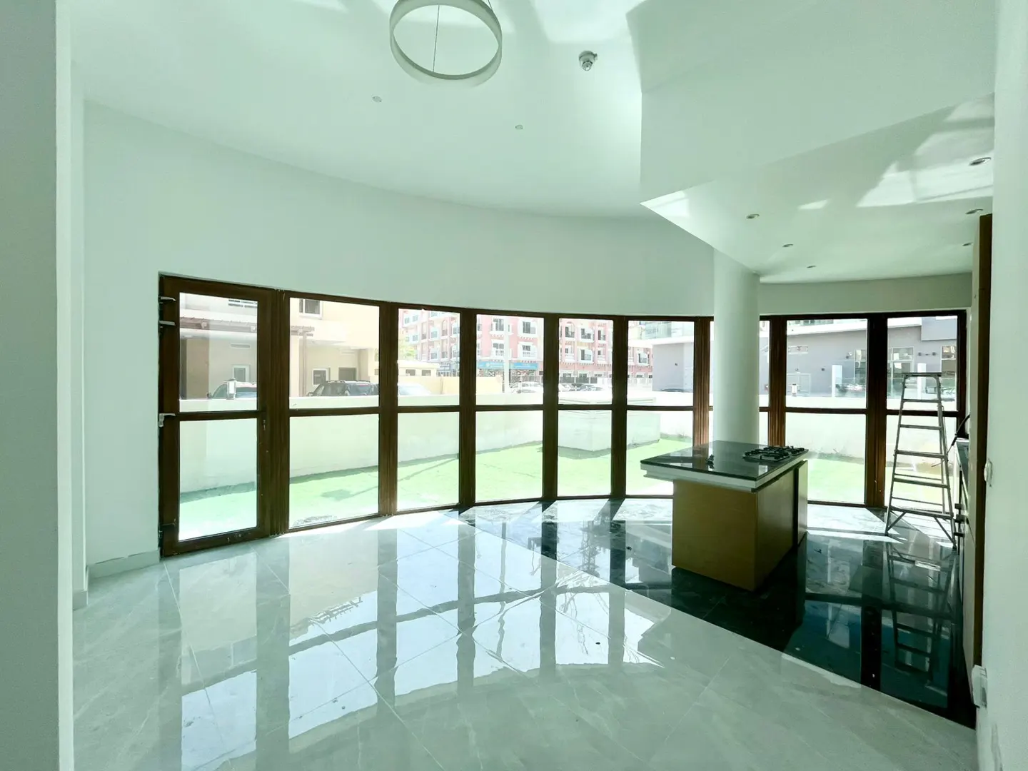 Bright, empty room with white walls, marble floors, and dark wood-framed windows. A kitchen island and ladder are visible.