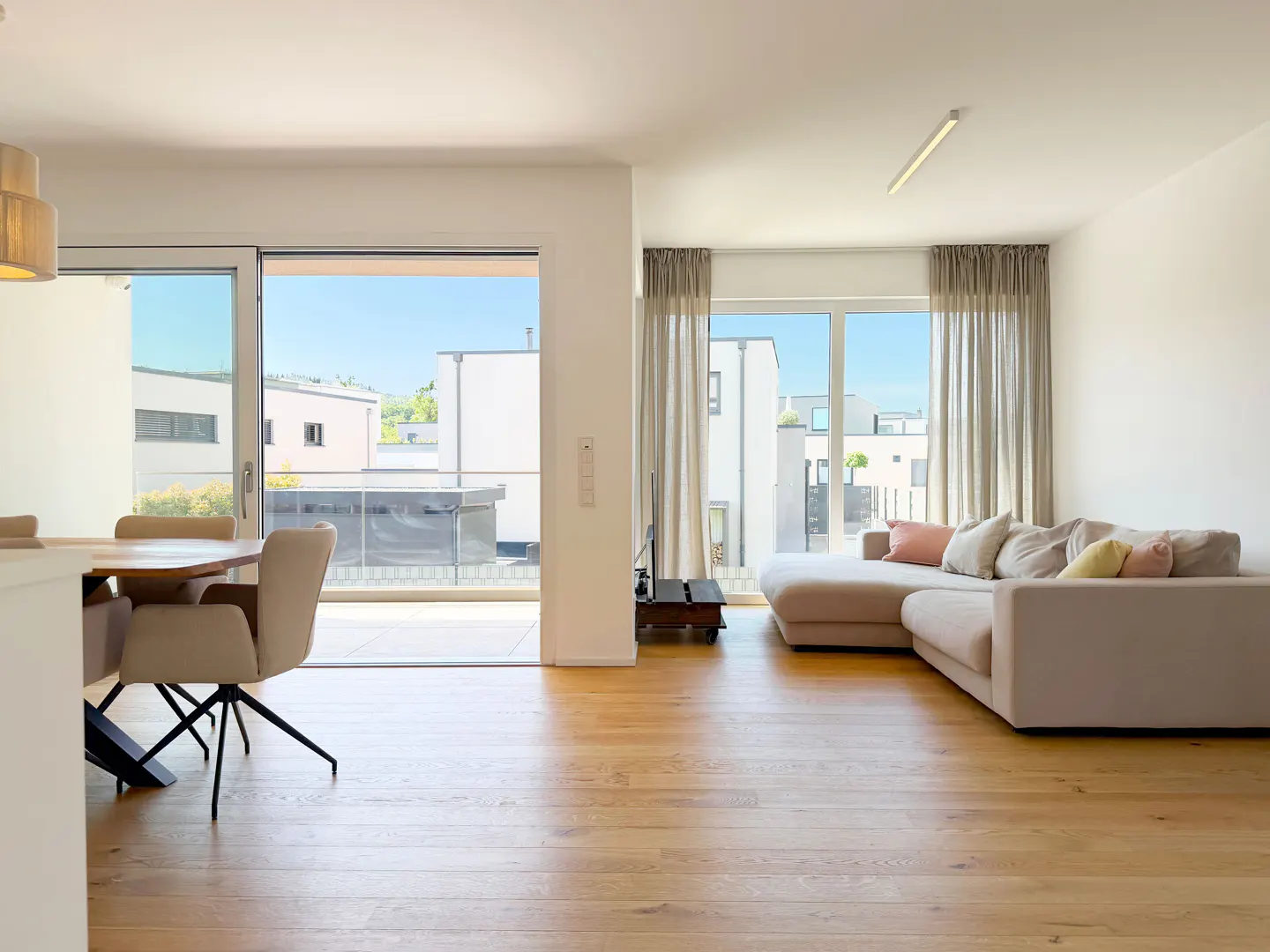 Bright open-plan living room with light wood floors, a beige sectional sofa, dining area, and large windows to a balcony.