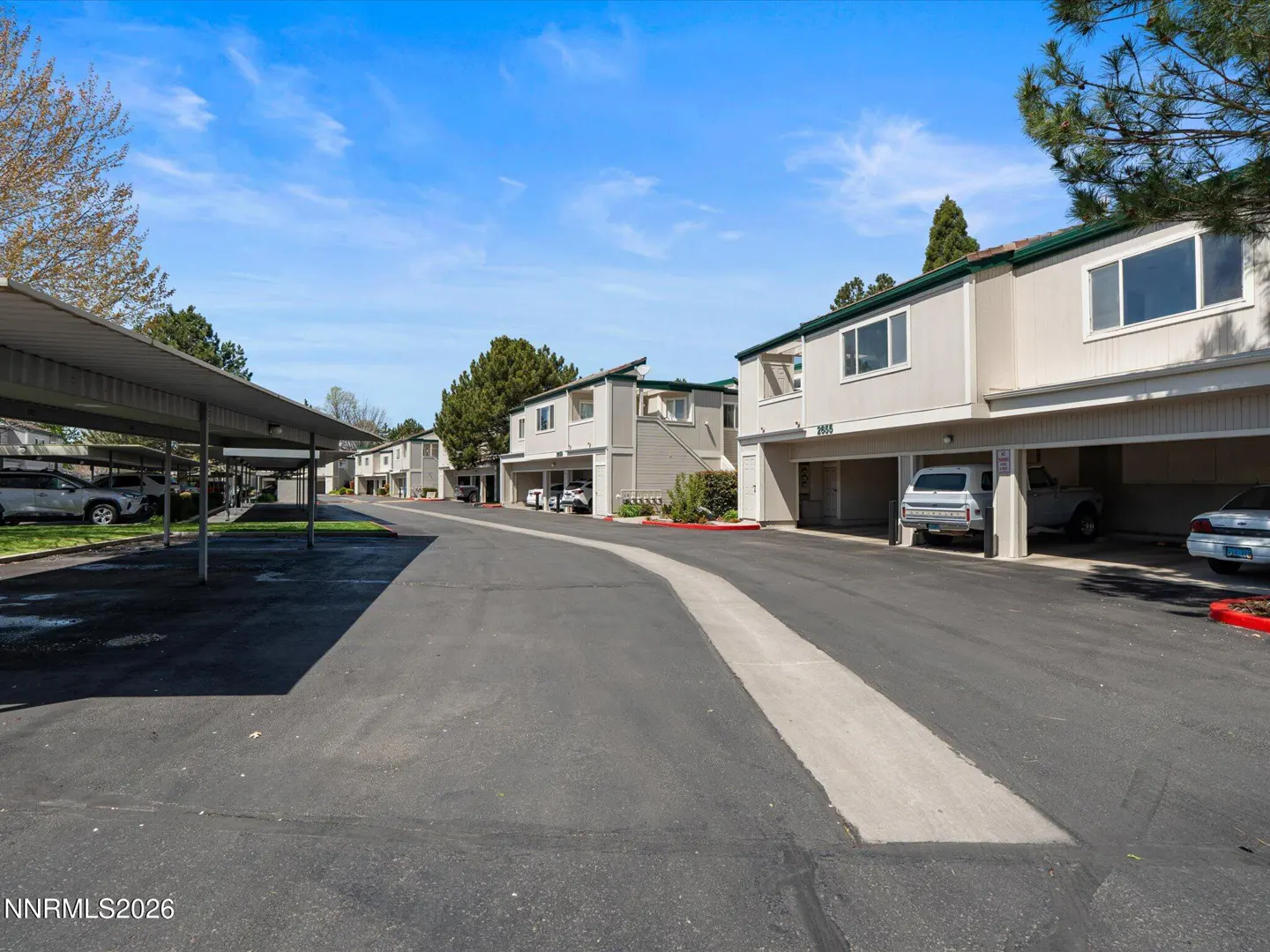 Exterior view of a row of two-story, beige apartments with carports and parked cars under a blue sky.