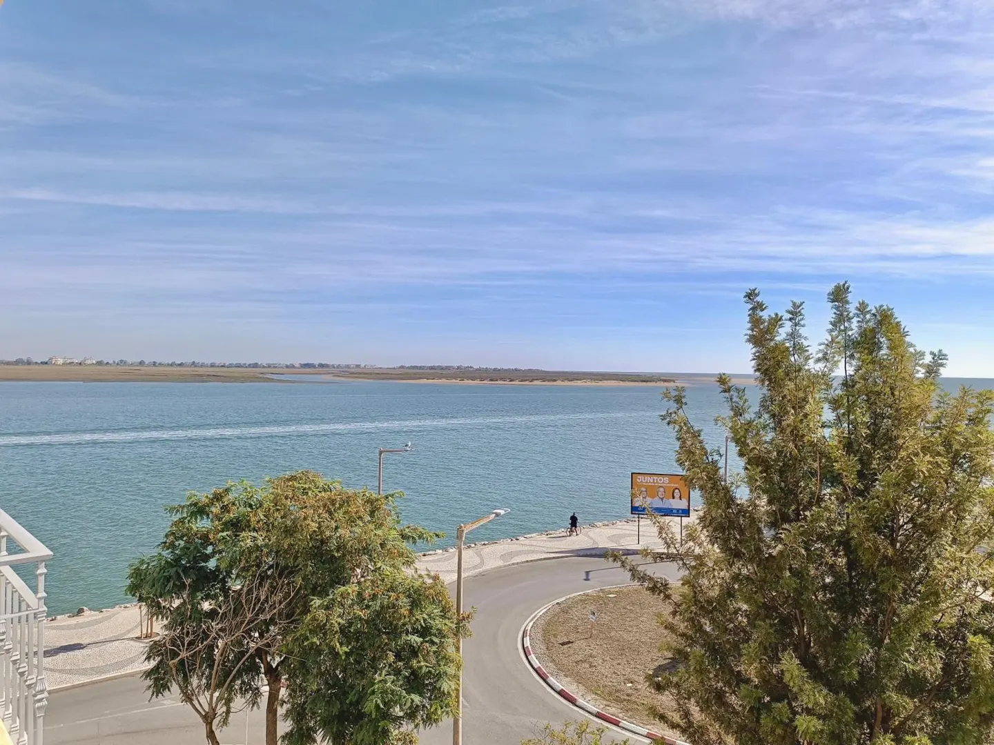View of a blue bay under a blue sky with trees in the foreground and a road with a person walking along the water.