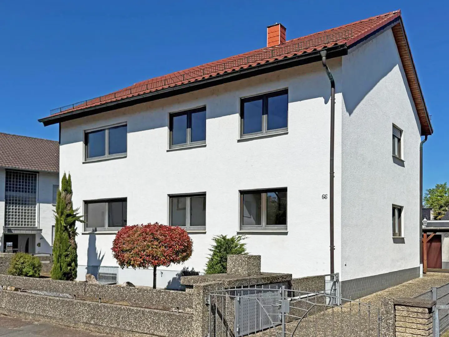 Two-story white house with a red tile roof and dark framed windows under a clear blue sky. A small red tree sits in front.