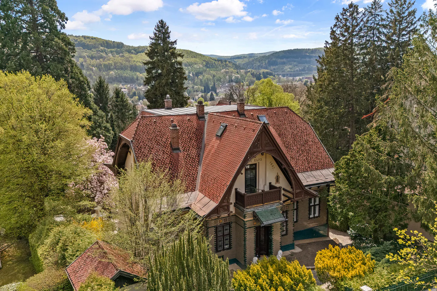 Exterior view of a large, two-story house with a red tile roof, surrounded by lush green trees and a distant mountain range.