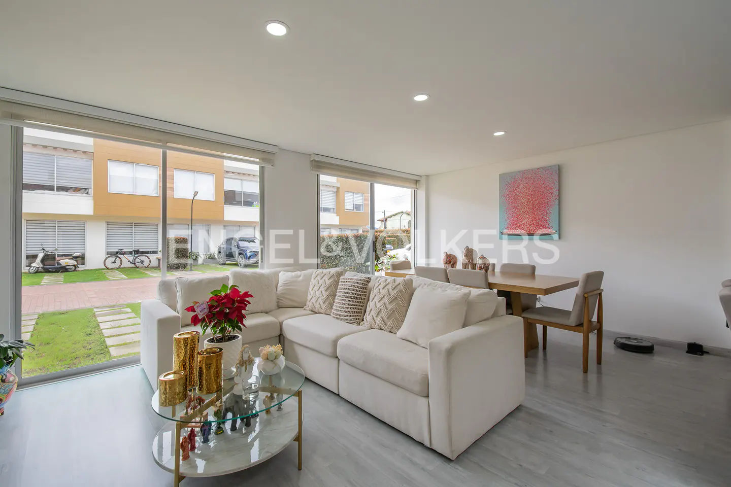 Bright living room with a white sectional sofa, glass coffee table, and dining table. Large windows overlook a green lawn and neighboring houses.