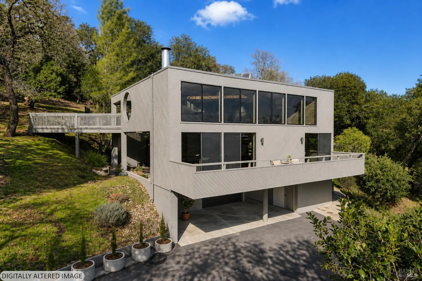 Exterior view of a modern gray house with large windows, a deck, and a carport surrounded by trees.