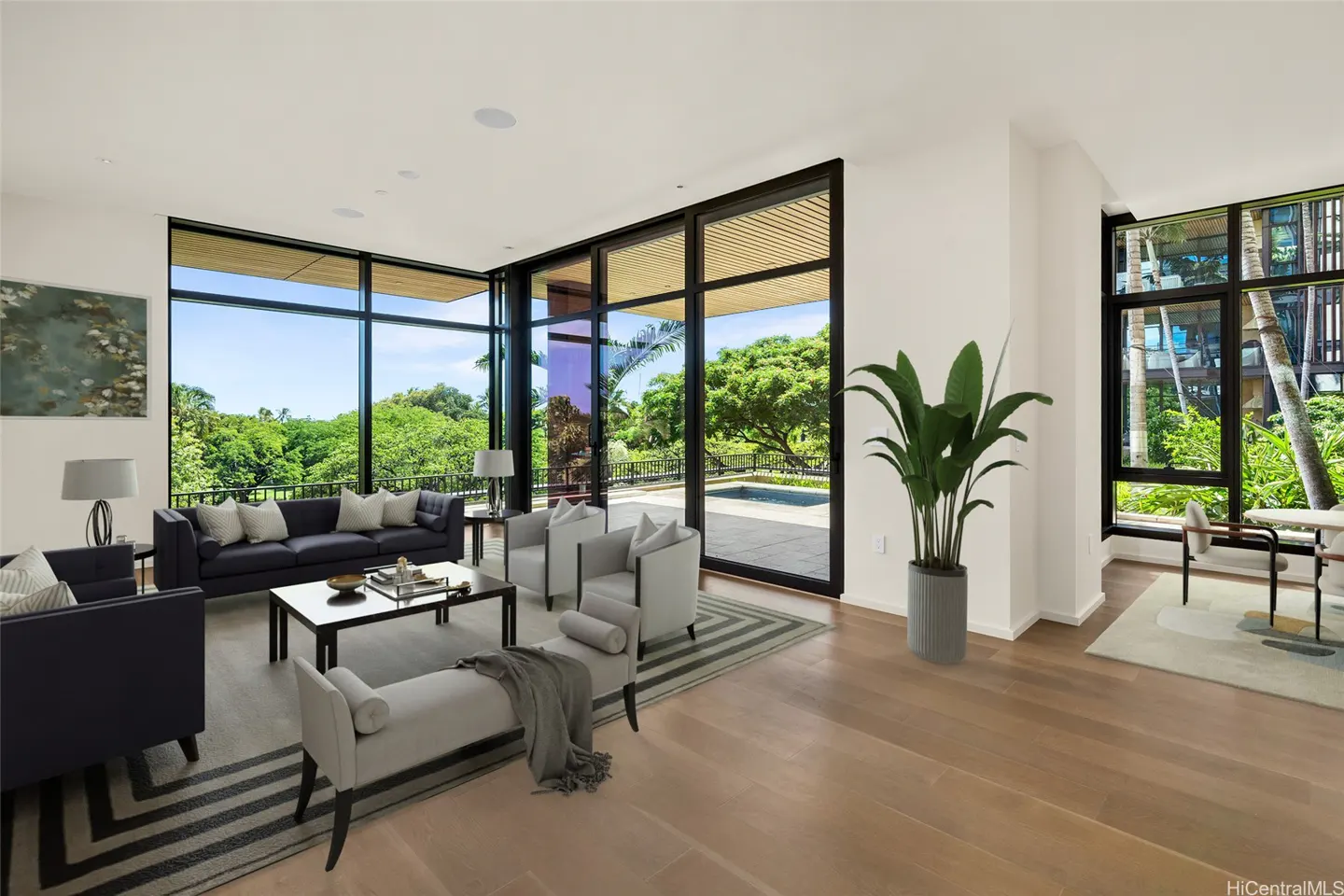 Bright living room with hardwood floors, black sofas, and gray chairs. Large windows offer views of lush greenery and a pool. A potted plant adds a touch of nature.