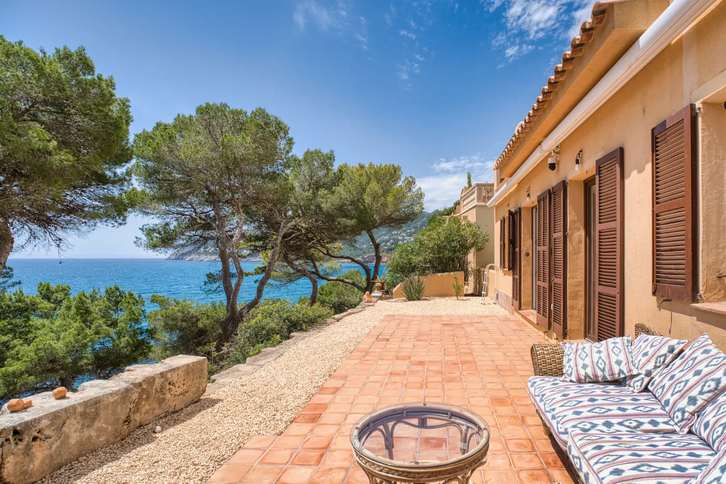 Outdoor patio with terracotta tiles, a patterned sofa, and a glass table. Ocean view with trees and blue sky.