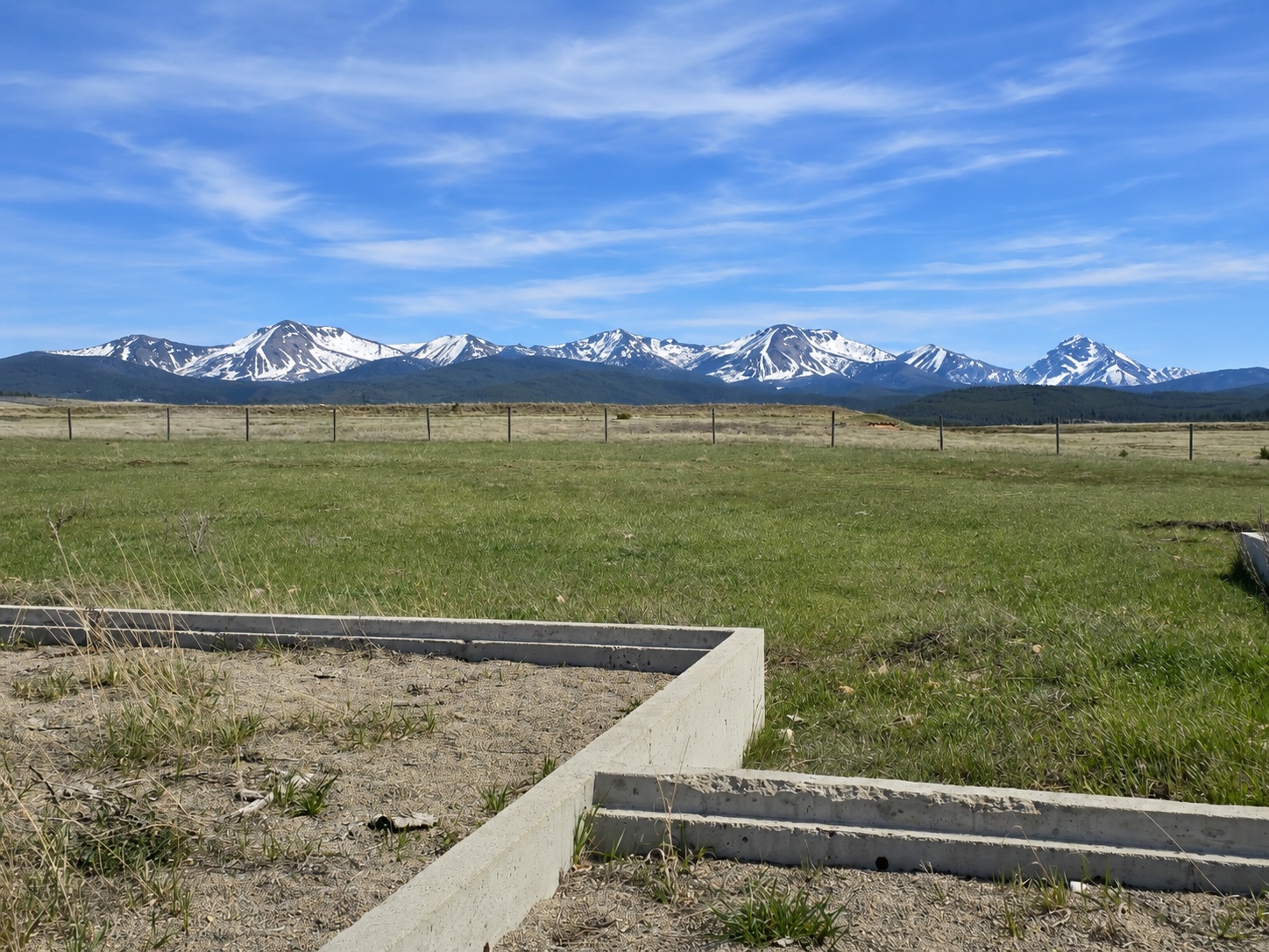 A grassy lot with a concrete foundation under a blue sky with snow-capped mountains in the distance.