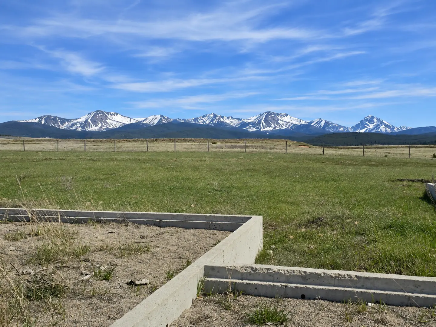A grassy lot with a concrete foundation under a blue sky with snow-capped mountains in the distance.
