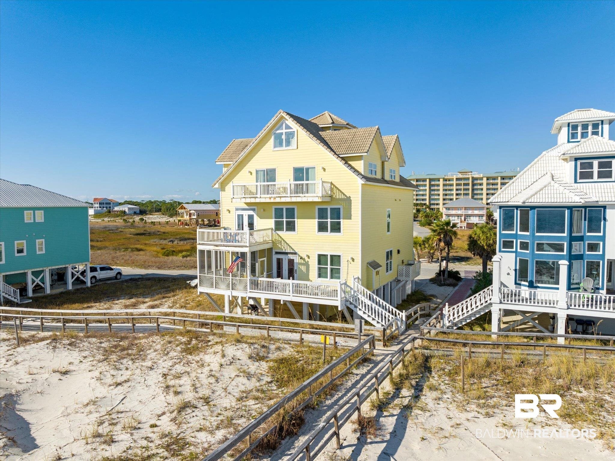 A three-story yellow beach house with white balconies, situated on a sandy dune with a wooden walkway under a clear blue sky.
