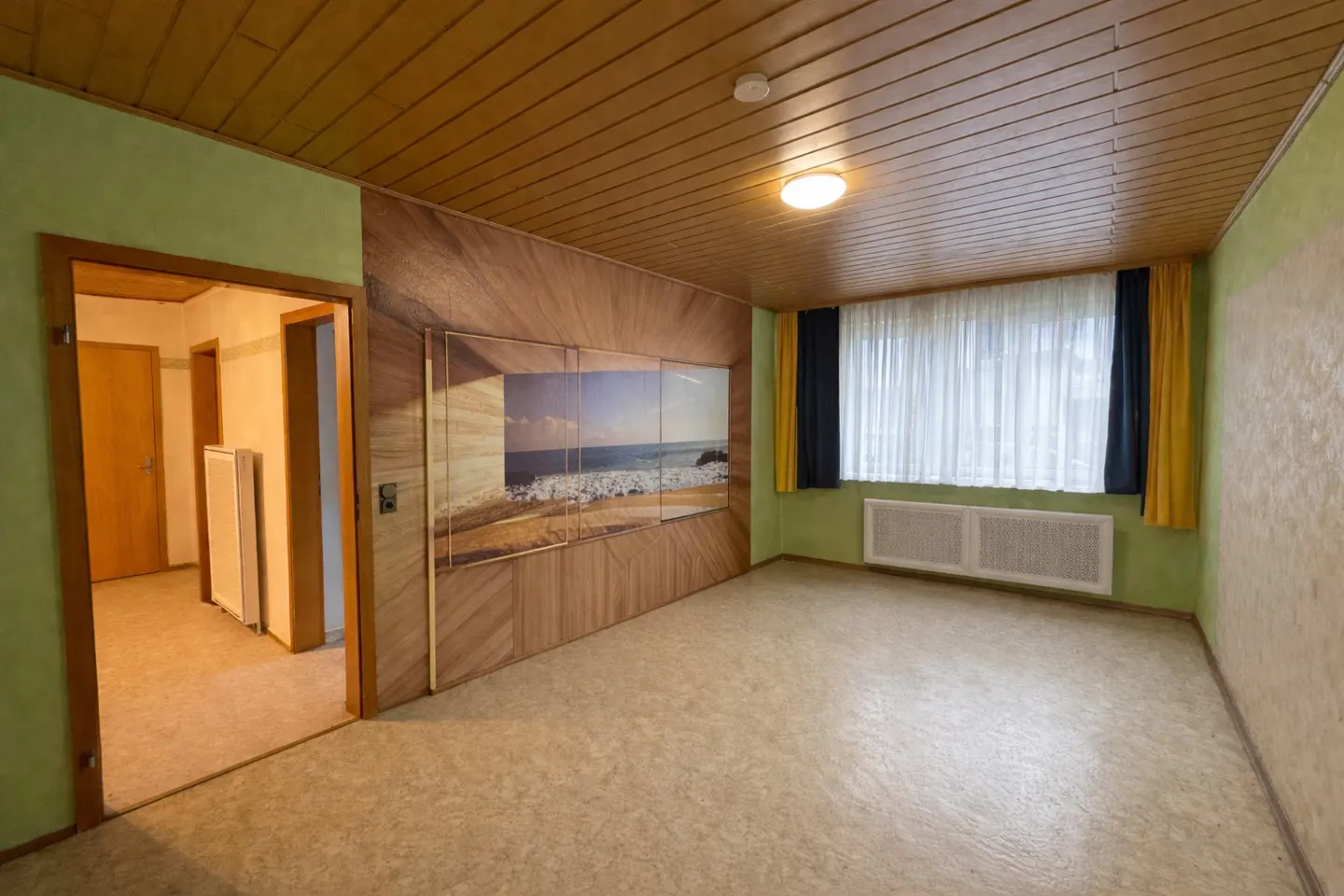 A room with wood-paneled walls, a beach mural, and a window with blue and yellow curtains.