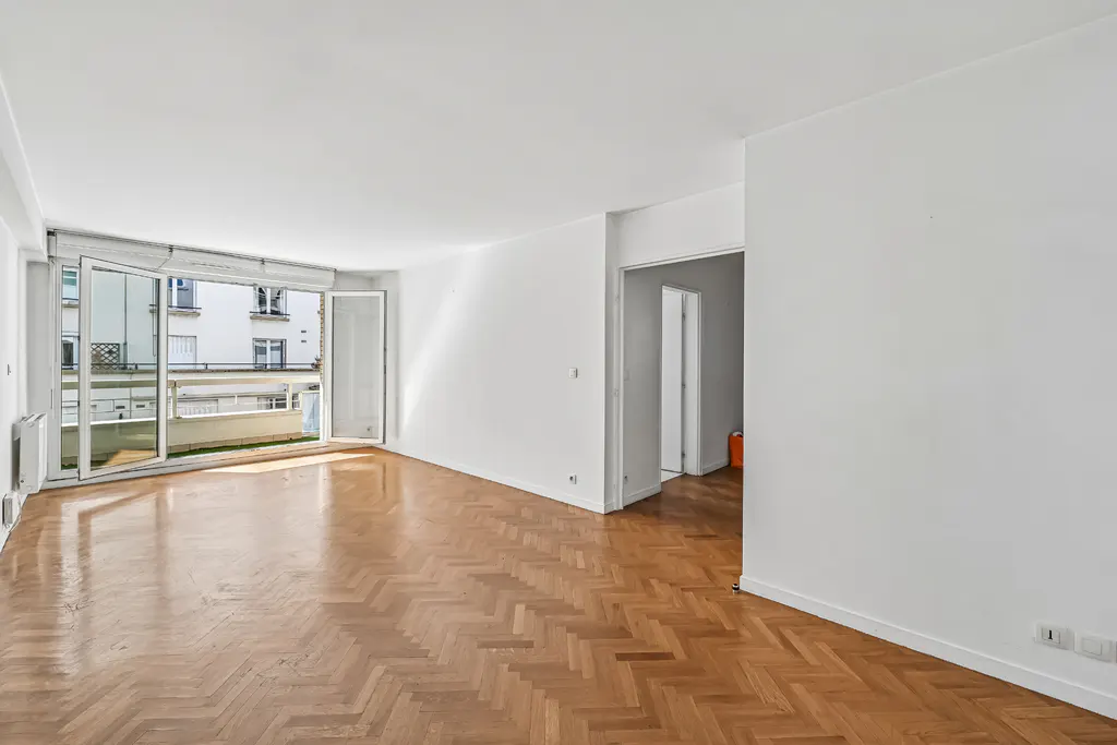 Bright, empty room with herringbone wood floor, white walls, and open balcony doors. View of building exterior through the doors.