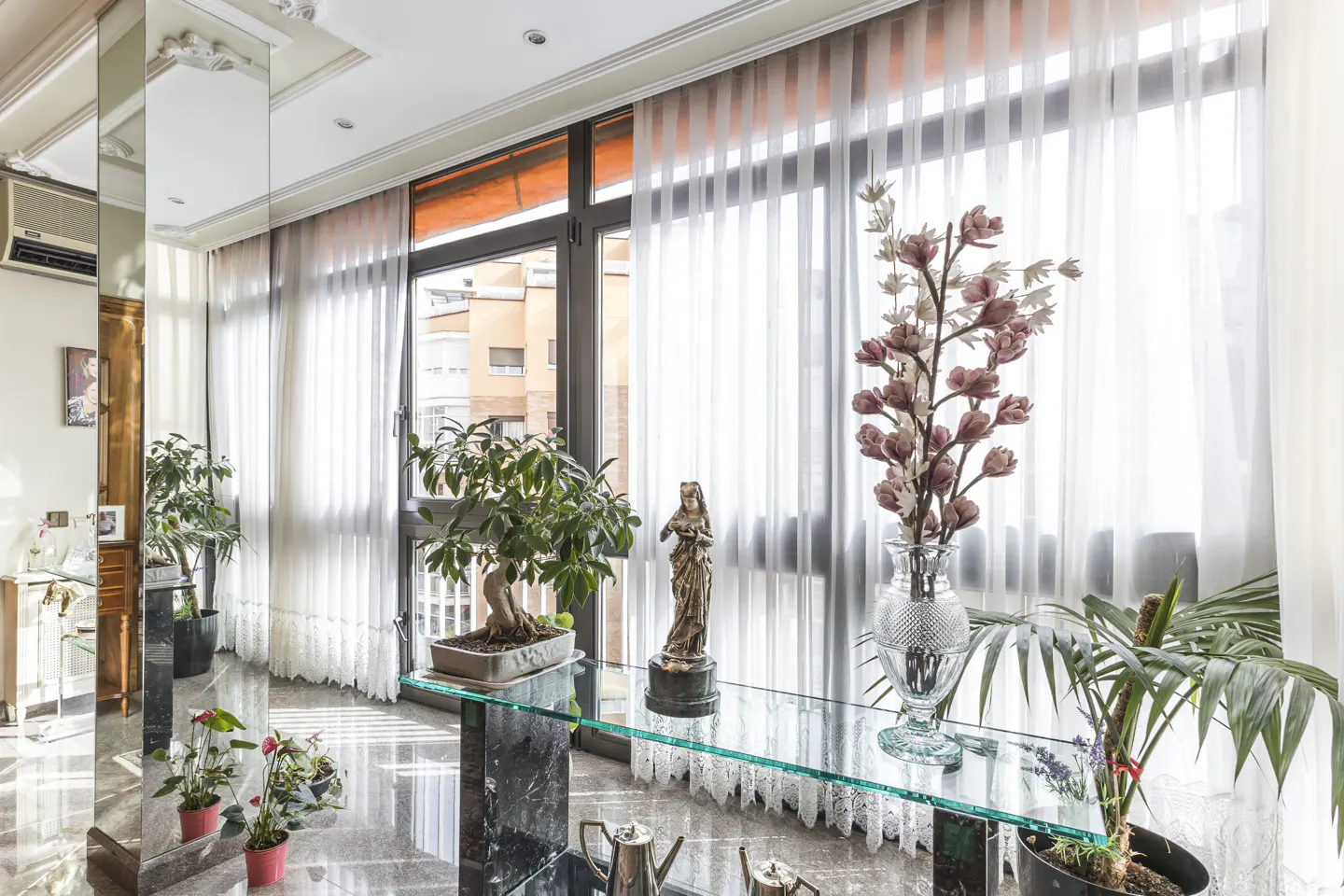 Living room with marble floors, sheer curtains, and a glass table. Plants, a statue, and a vase of flowers decorate the table.