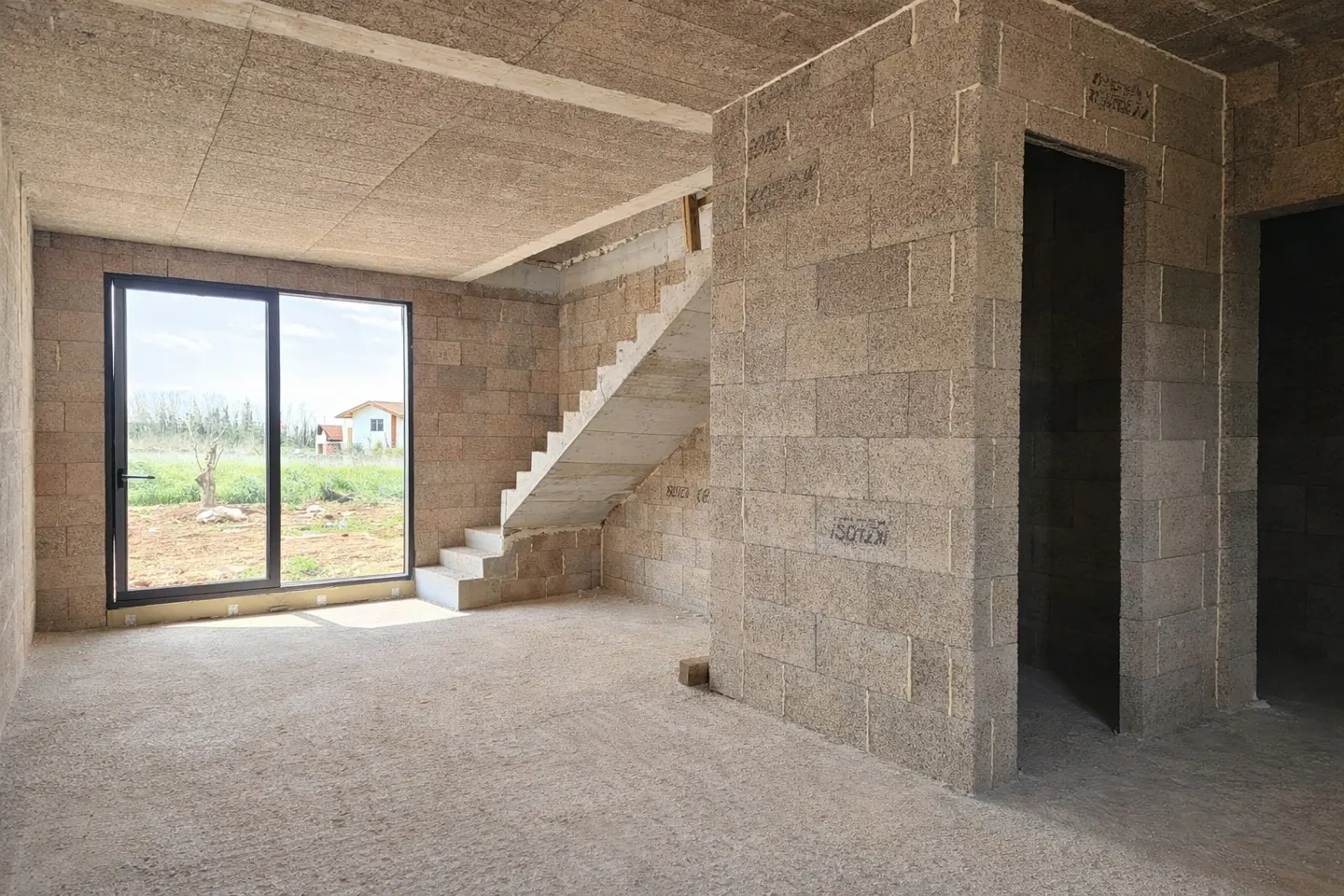 Unfinished room with concrete block walls, stairs, and a black-framed window showing a yard and house.