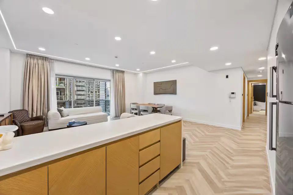 Bright, modern apartment interior with white walls, herringbone wood floors, and a kitchen island. A dining table and living area are visible.