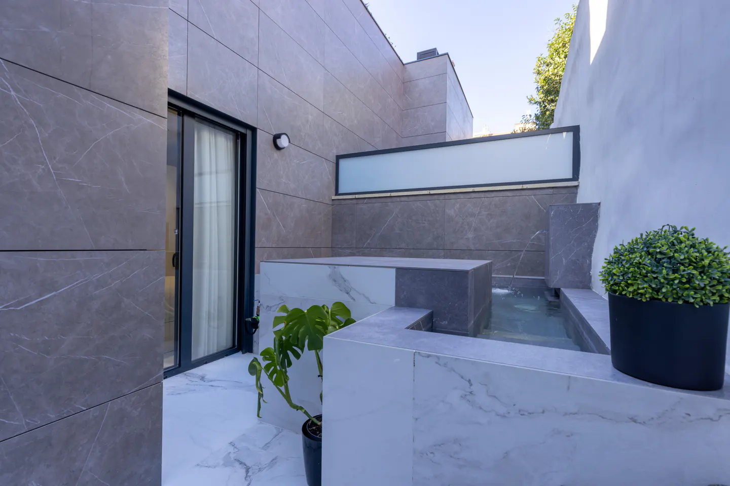Modern patio with gray marble walls, a small pool with a fountain, and potted plants. A sliding glass door leads indoors.