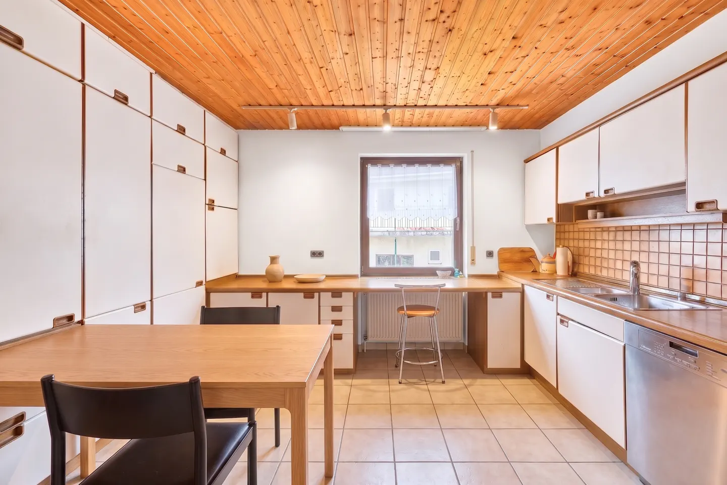 A bright kitchen with white cabinets, a wooden ceiling, and a table with chairs. A window is visible in the background.