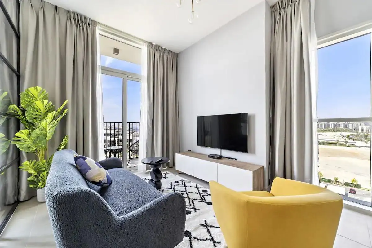 Bright living room with blue sofa, yellow chair, TV, and balcony view. Neutral curtains frame the windows. A patterned rug adds texture to the space.