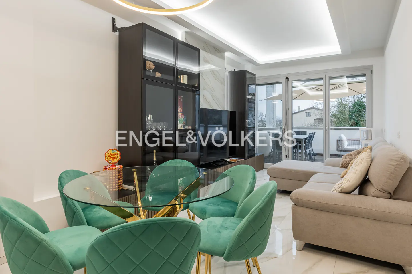 Bright living room with a glass table, green velvet chairs, and a beige sofa leading to a balcony. Dark cabinets and a TV stand against a marble wall.