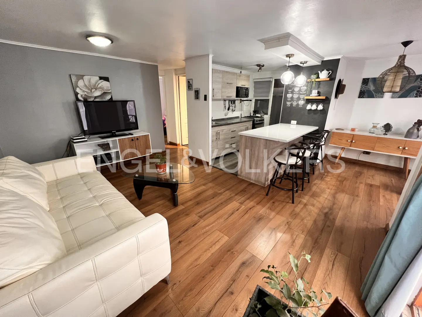 Open-concept living space with wood floors, a white sofa, TV, kitchen island with stools, and modern decor.