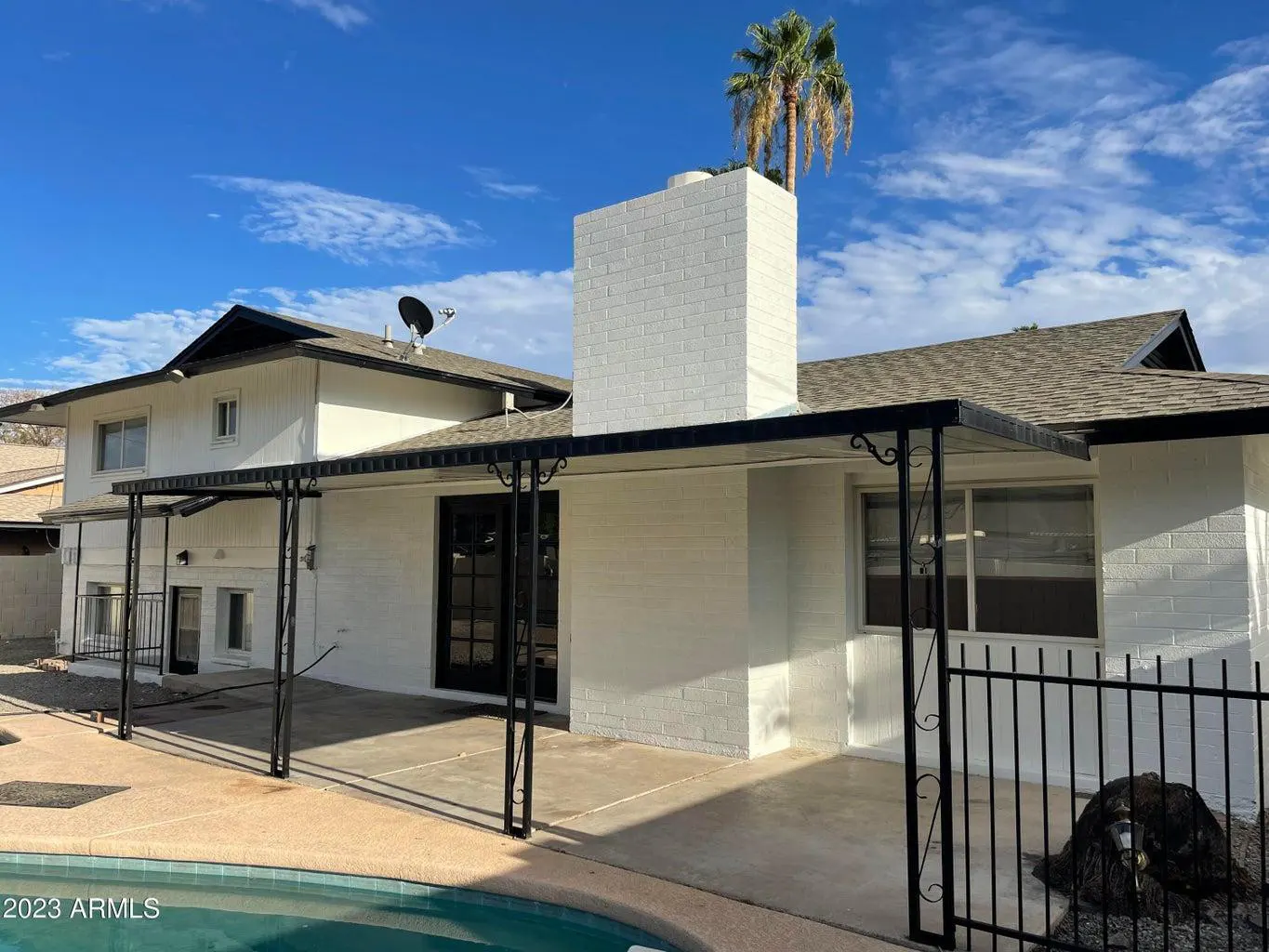 Exterior view of a white two-story house with a pool, patio cover, and a tall white brick chimney. A palm tree is visible behind the chimney.