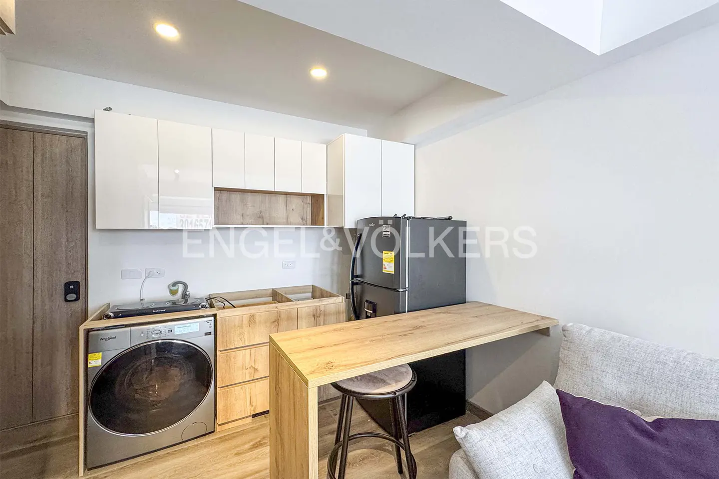 A modern apartment interior shows a washer, cabinets, fridge, and a small table with a stool. A gray sofa is visible on the right.