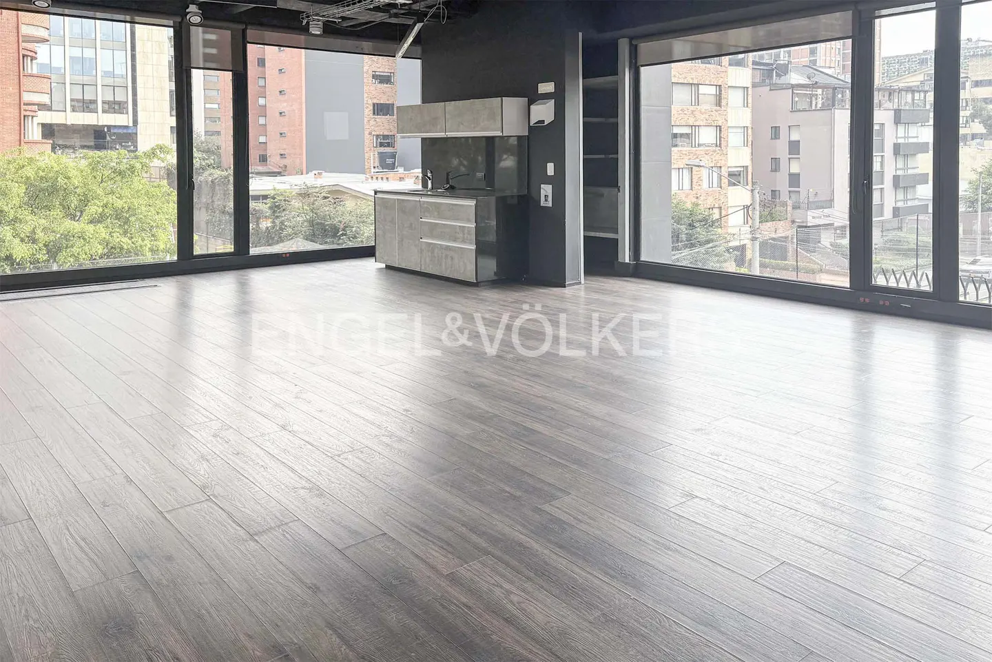 Empty room with dark wood floors, floor-to-ceiling windows, and a small gray kitchen area. City buildings are visible through the windows.