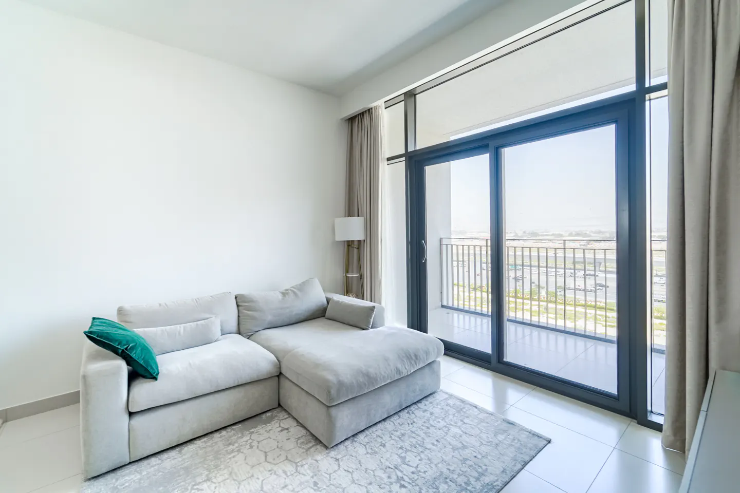 Bright living room with a gray sectional sofa, a green pillow, and a patterned rug. Sliding glass doors lead to a balcony with a city view.