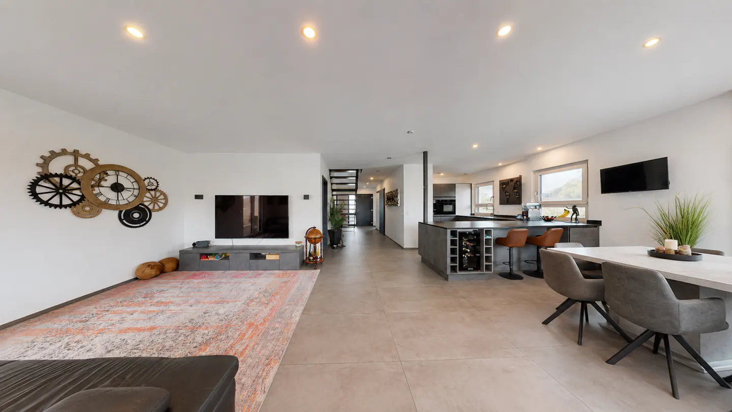Open-plan living area with a large gear-themed wall clock, grey dining set, kitchen island, and a patterned area rug.