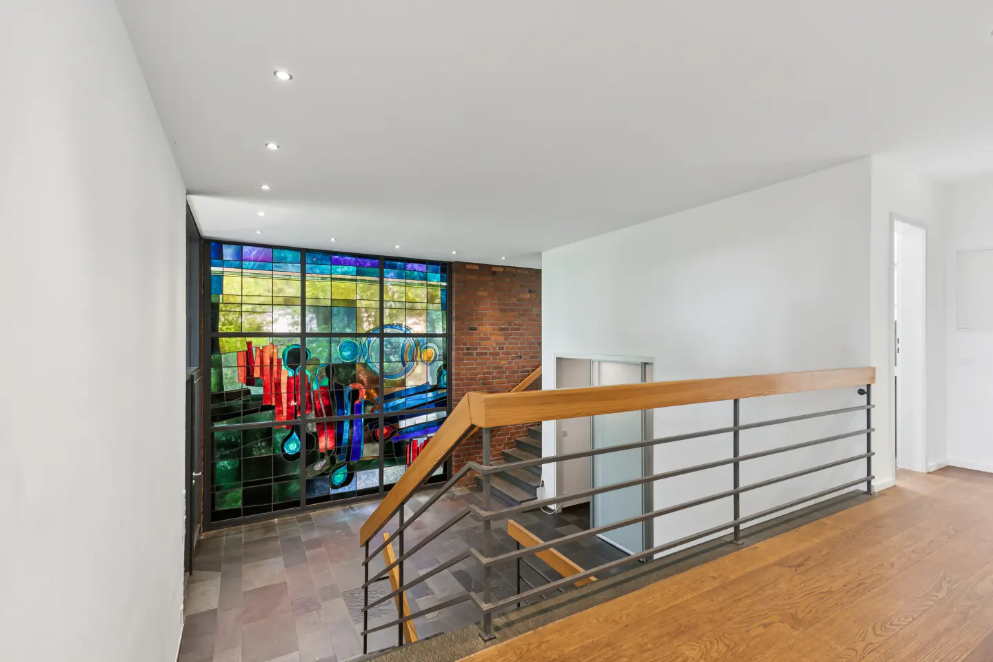 Modern staircase with wooden handrail, metal railings, and a large, colorful stained-glass window against a brick wall.