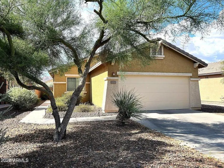 A tan single-story home with a two-car garage and a brown roof. A tree is in the front yard.