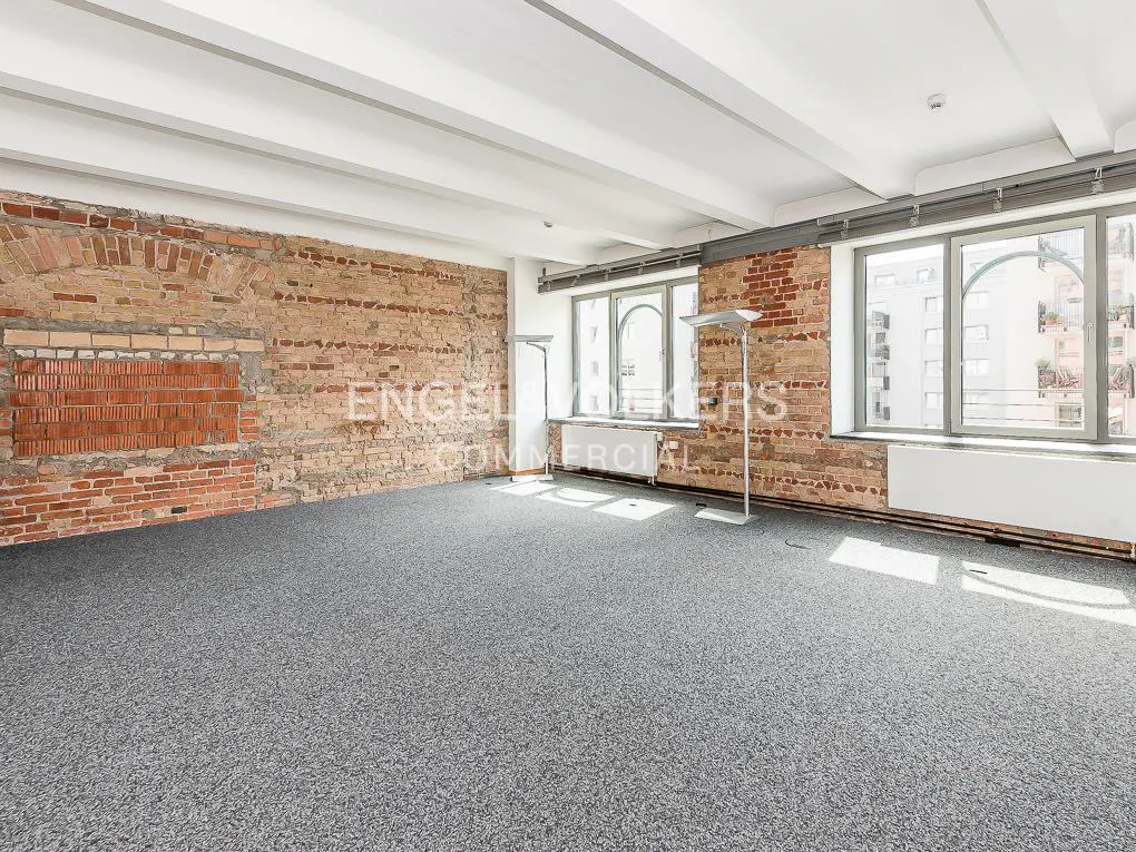 Empty room with exposed brick walls, gray carpet, white ceiling beams, and large windows.