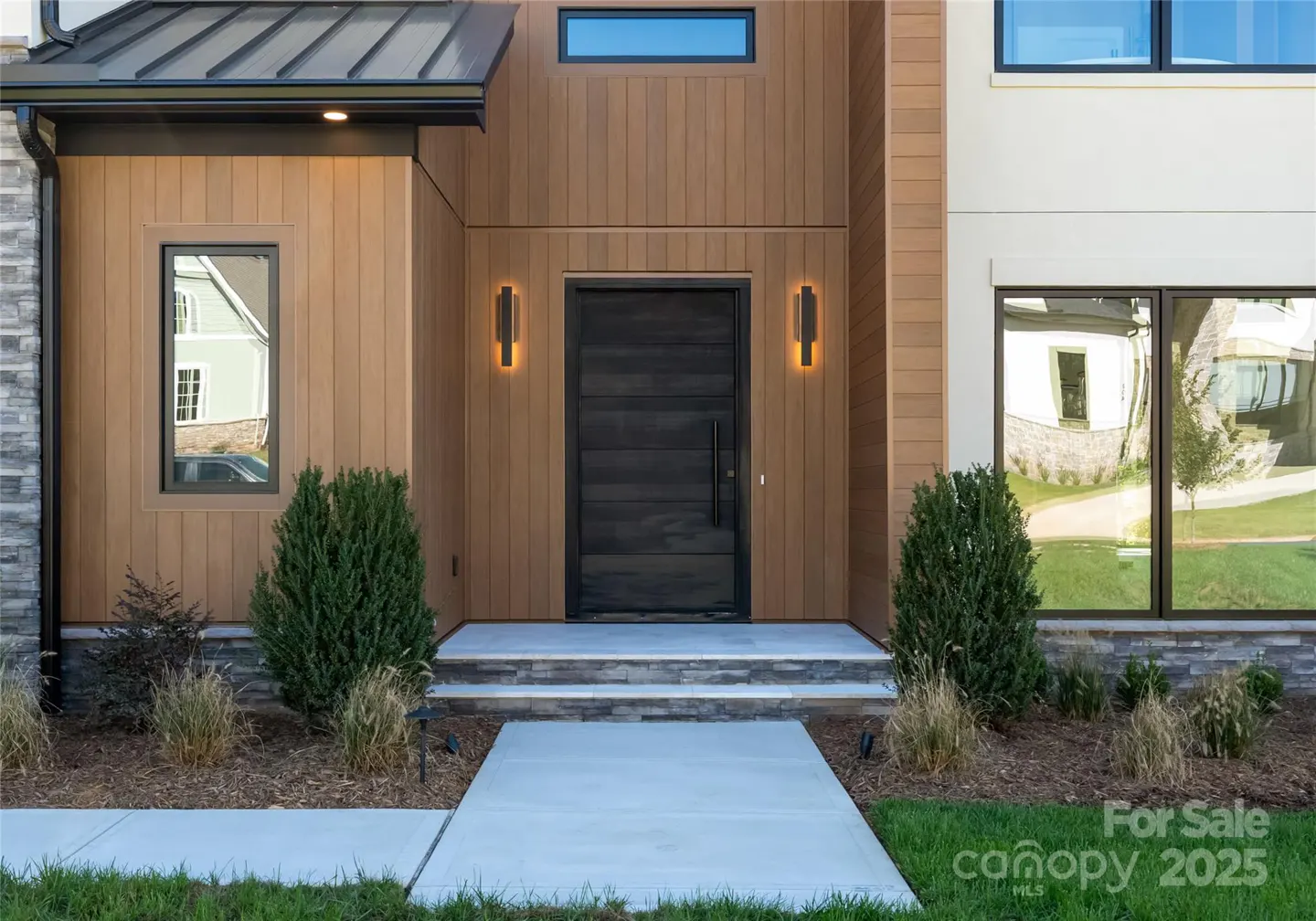 Modern home exterior with a dark wood door, tan siding, and stone accents. A concrete walkway leads to the entrance.
