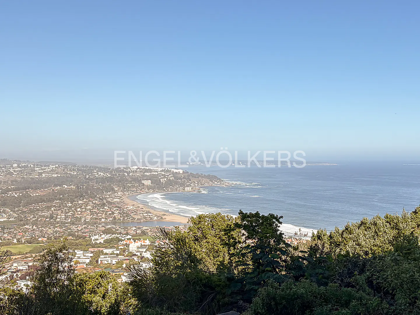Scenic view of a coastal city with a sandy beach, blue ocean, and green trees under a clear sky.