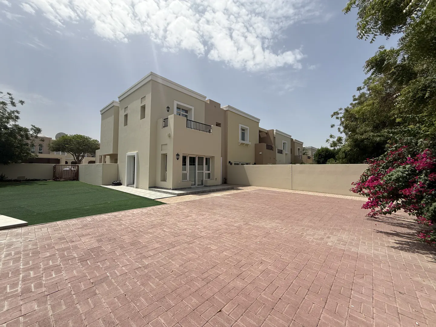 Exterior view of a two-story beige house with a balcony, a red brick driveway, and a green lawn under a blue sky.