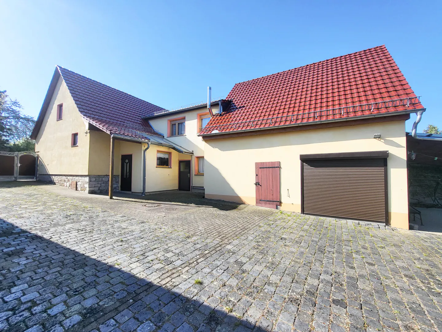 Exterior view of a yellow house with a red tile roof and a cobblestone driveway on a sunny day.
