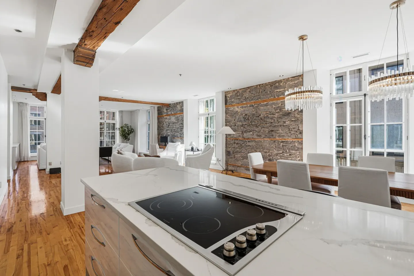 Open-concept living space with white walls, wood floors, exposed beams, and a stone accent wall. A kitchen island with a cooktop is in the foreground.