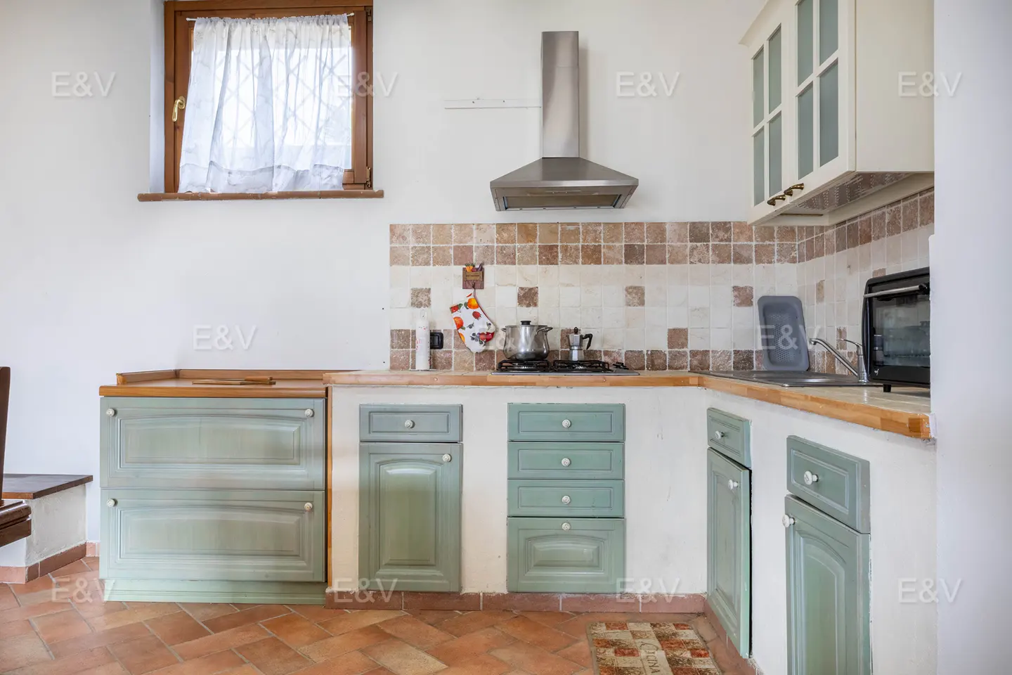 A kitchen with light green cabinets, a stainless steel range hood, and a window with white curtains.
