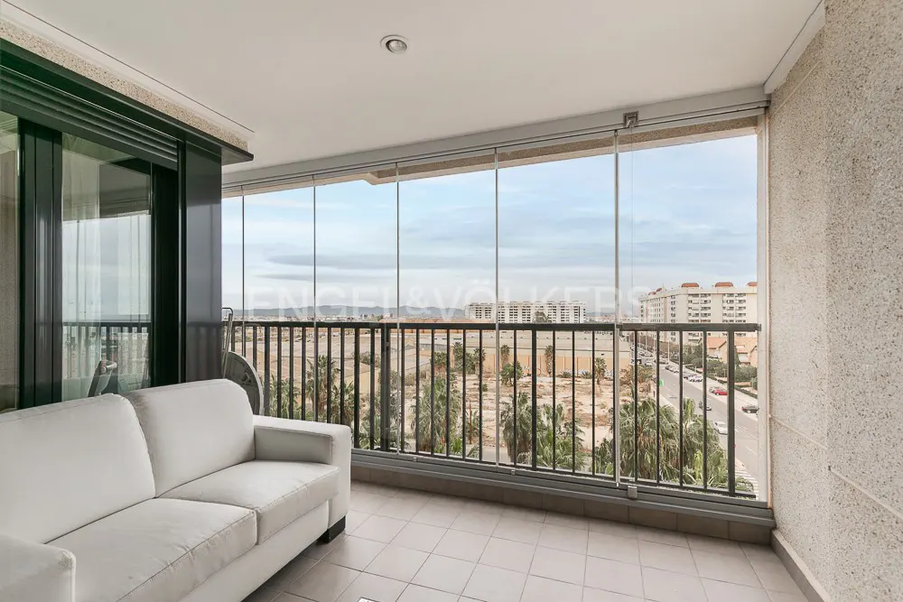 A white sofa on a tiled balcony enclosed by glass panels, overlooking a city street with palm trees and distant buildings.